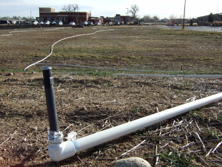 Black sprinkler body attached by an elbow to white lateral irrigation piping laid above ground before installation.